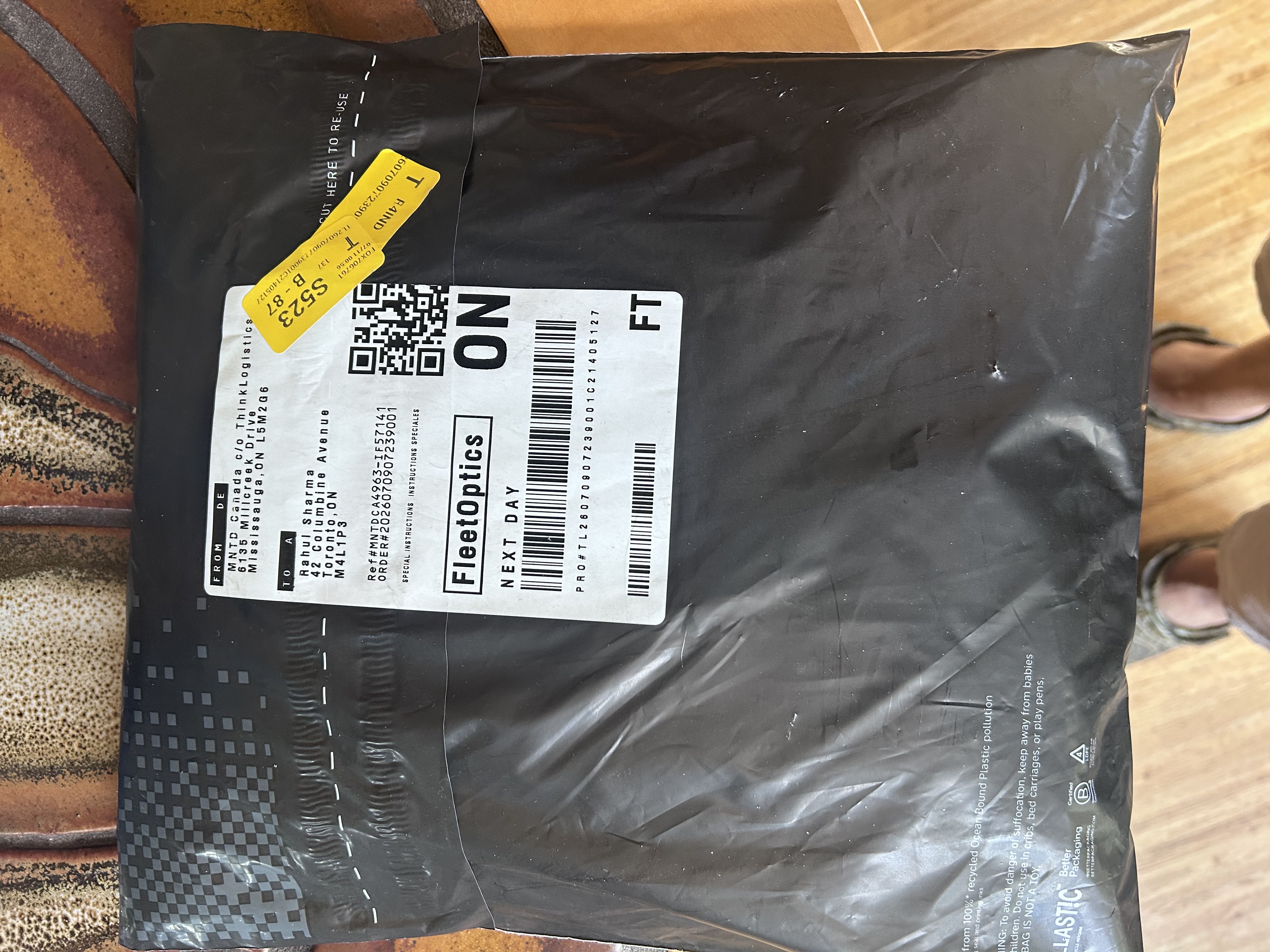 MNTD FleetOptics polybag received at doorstep · black recycled ocean-bound plastic bag with shipping label showing FleetOptics Next Day service, MNTD wordmark visible